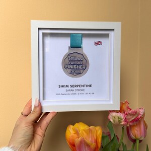 Puede incluir: Una medalla de plata con una cinta azul, enmarcada en blanco. La medalla dice "Swim Serpentine Two Mile Finisher" con una bandera del Reino Unido en la esquina. El texto debajo de la medalla dice "Swim Serpentine Sarah Stroke 20th September 2025 | 2 miles | 01:42:58"