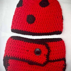 Może przedstawiać: Ręcznie robiony szydełkowy strój dla dziecka w kształcie biedronki. Zestaw zawiera czerwony kapelusz z czarnymi kropkami i pasującą pieluchę. Krawędzie są obszyte na czarno, a pielucha ma zapięcie na guzik. Uroczy kostium dla dziecka.