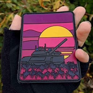 Peut inclure: Un écusson rectangulaire avec un motif de char d'assaut sur fond de coucher de soleil. Le patch présente un char noir au-dessus de plantes roses et violettes. Le fond passe du violet au rose et au jaune, avec un grand soleil jaune.
