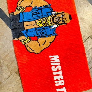 May include: Red rug featuring a cartoon image of a muscular man wearing a blue denim vest and gold chains. The text "Mister T" is printed in white on the rug.