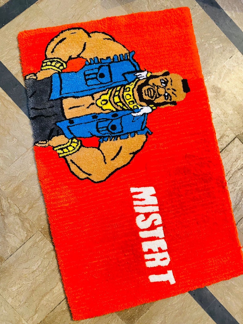 May include: Red rug featuring a cartoon image of a muscular man wearing a blue denim vest and gold chains. The text "Mister T" is printed in white on the rug.