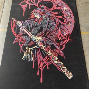 May include: A black rug featuring a red and pink design of a figure holding a sword. The figure has long red hair and is standing in front of a large, red, crescent moon.