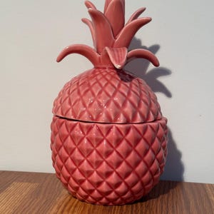 May include: A pink ceramic pineapple-shaped container with a lid. The container has a textured diamond pattern and a leafy top. The pineapple is sitting on a wooden surface.