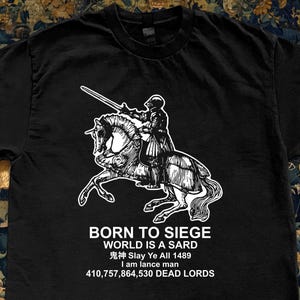 May include: Black t-shirt featuring a white graphic of a knight on horseback with a sword. Text below reads "BORN TO SIEGE WORLD IS A SARD 鬼神 Slay Ye All 1489 I am lance man 410,757,864,530 DEAD LORDS."