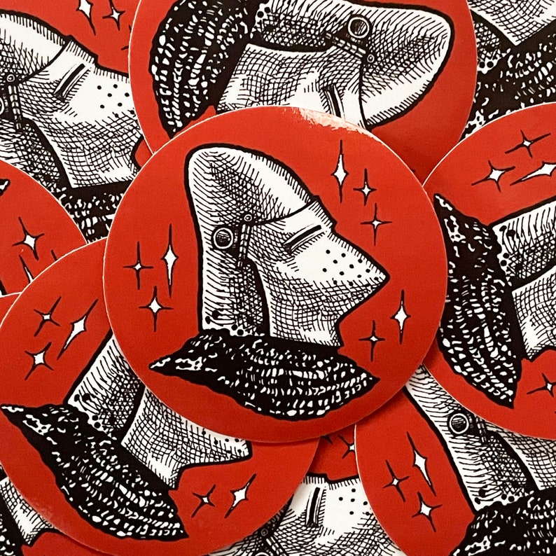 May include: A set of red and black circular stickers featuring a knight's helmet with a detailed design. The helmet is surrounded by a white background with small stars.