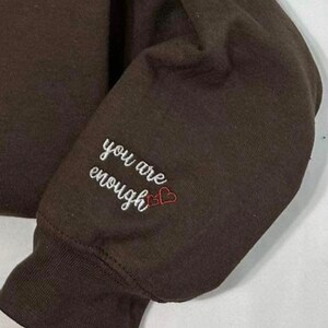 May include: A brown sweatshirt with the phrase "you are enough" embroidered in white with two red hearts.