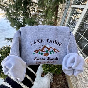May include: Gray sweatshirt with embroidered text "LAKE TAHOE" and a mountain scene with a bear. The text "Sierra Nevada CALIFORNIA" is also embroidered below the mountain scene.