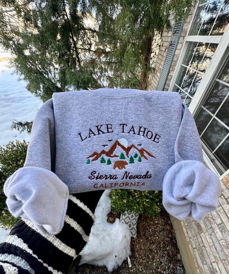 May include: Gray sweatshirt with embroidered text "LAKE TAHOE" and a mountain scene with a bear. The text "Sierra Nevada CALIFORNIA" is also embroidered below the mountain scene.