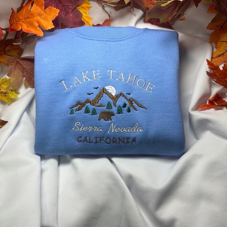 May include: Light blue sweatshirt with embroidered text "LAKE TAHOE" and a mountain scene with a bear. The text "Sierra Nevada CALIFORNIA" is also embroidered on the sweatshirt.