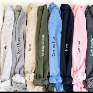May include: A collection of folded sweatshirts in various colors, including white, ash, sport grey, sand, military green, Carolina blue, navy, soft pink, dark heather, and black. Each sweatshirt has its color name printed on it.