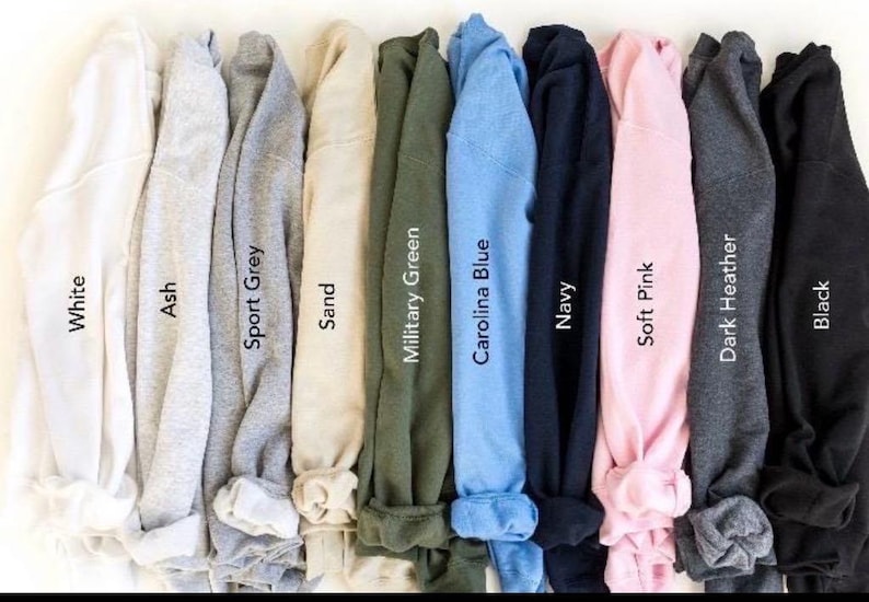May include: A collection of folded sweatshirts in various colors, including white, ash, sport grey, sand, military green, Carolina blue, navy, soft pink, dark heather, and black. Each sweatshirt has its color name printed on it.