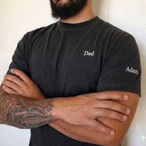 May include: Dark gray t-shirt with the word "Dad" printed on the chest and "Adam" on the sleeve. The shirt has a crew neck and short sleeves. The shirt is worn by a person with a beard and a tattoo on their arm.