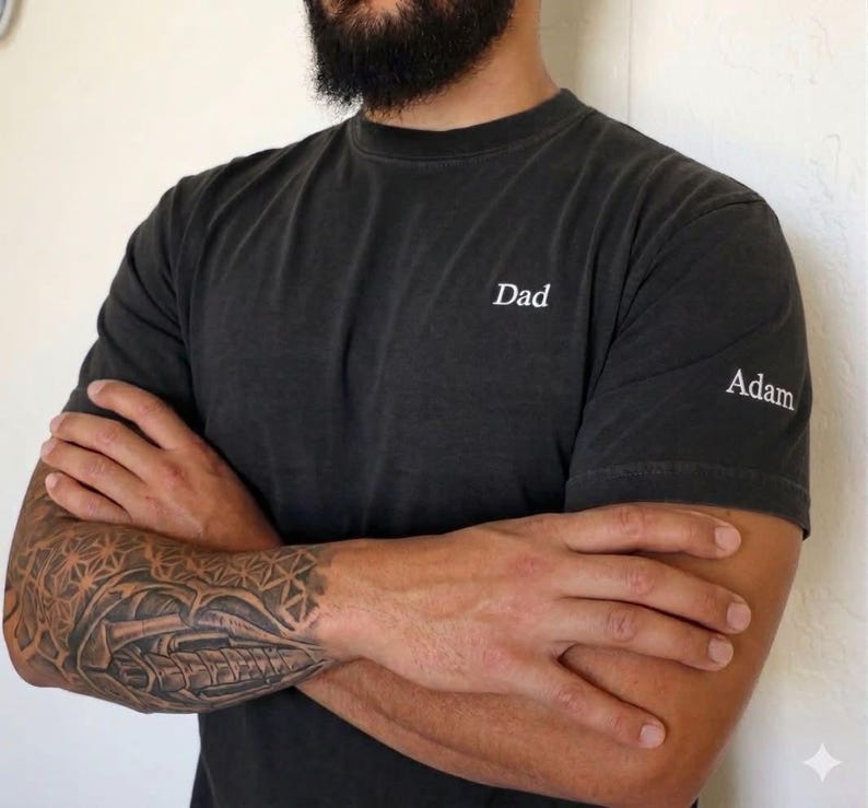 May include: Dark gray t-shirt with the word "Dad" printed on the chest and "Adam" on the sleeve. The shirt has a crew neck and short sleeves. The shirt is worn by a person with a beard and a tattoo on their arm.