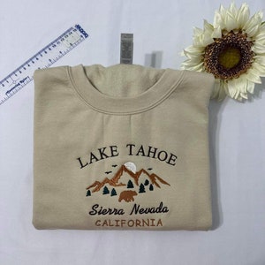 May include: A beige sweatshirt with an embroidered design of mountains, a bear, and trees. The text "LAKE TAHOE" is above the design, and the text "Sierra Nevada CALIFORNIA" is below the design.
