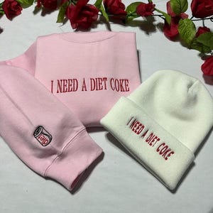 May include: A pink sweatshirt and a white beanie with the text "I need a diet coke" embroidered on both. The sweatshirt has a small embroidered image of a can of diet coke on the sleeve.