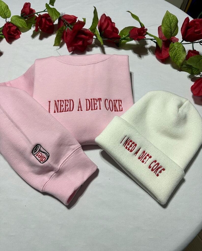 May include: A pink sweatshirt and a white beanie with the text "I need a diet coke" embroidered on both. The sweatshirt has a small embroidered image of a can of diet coke on the sleeve.