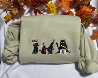 May include: A light green sweatshirt featuring an embroidered design of four black cats wearing Santa hats and reindeer antlers. The cats are standing in front of a Christmas tree.