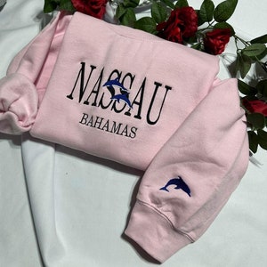 Puede incluir: Sudadera rosa con un diseño bordado en negro que dice "NASSAU BAHAMAS" con dos delfines. La sudadera tiene un delfín bordado en la manga.