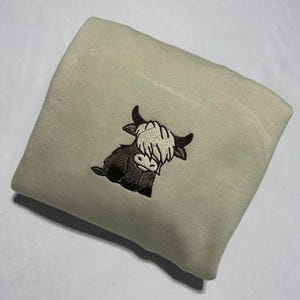 May include: A folded, light green sweatshirt featuring an embroidered Highland cow design. The cow is depicted in shades of brown and black with a cream-colored fringe. The sweatshirt is made of a soft material.