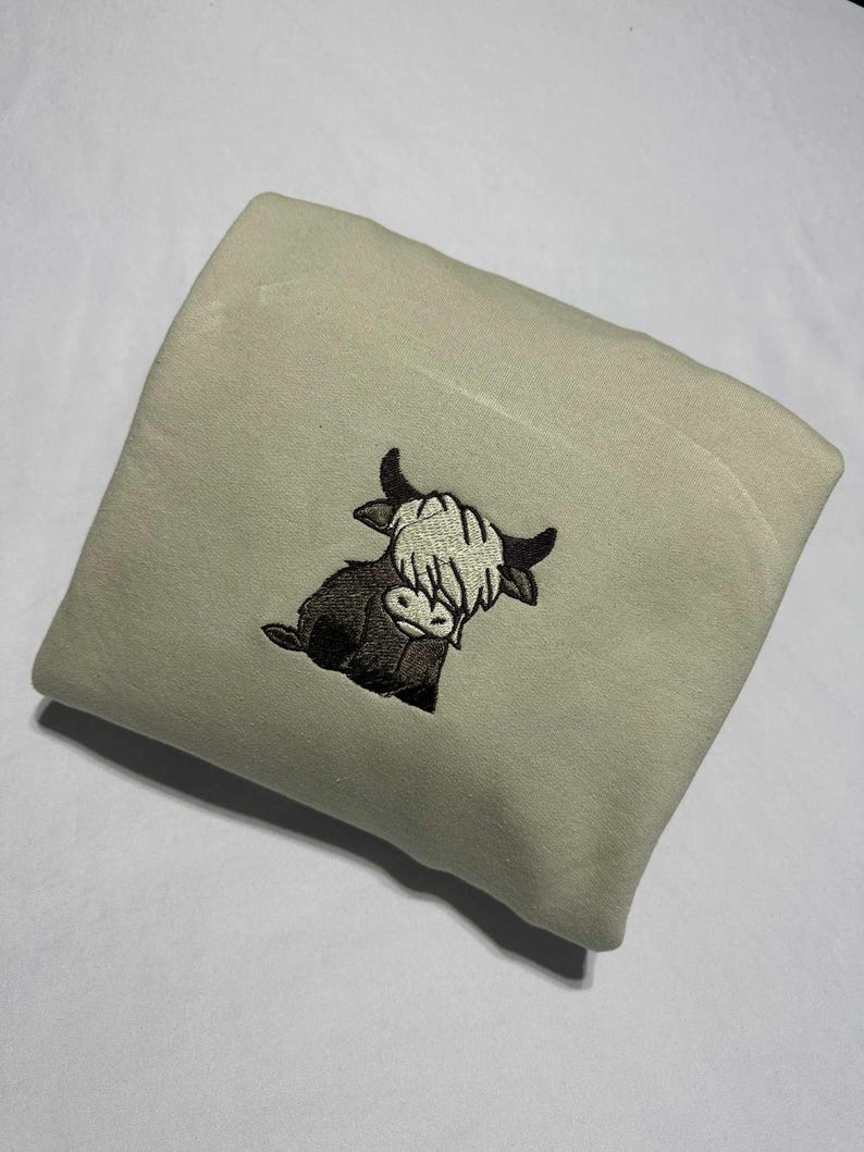May include: A folded, light green sweatshirt featuring an embroidered Highland cow design. The cow is depicted in shades of brown and black with a cream-colored fringe. The sweatshirt is made of a soft material.