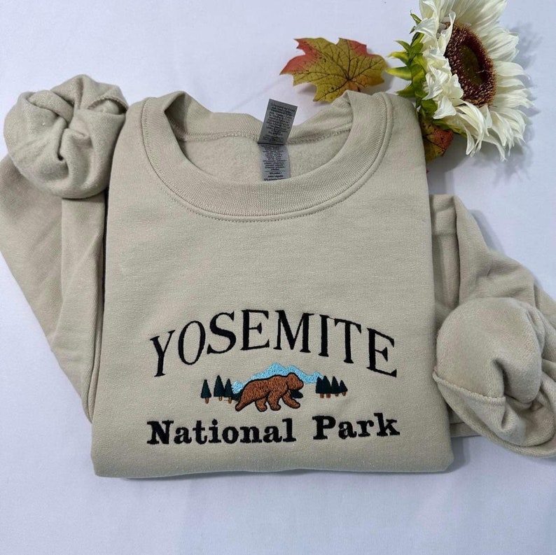 May include: A beige sweatshirt with the text "Yosemite National Park" embroidered in black. A brown bear with blue sky and green trees are embroidered below the text.