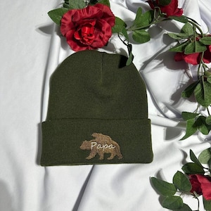 May include: A green knitted beanie with a brown bear silhouette and the word "Papa" embroidered in white.