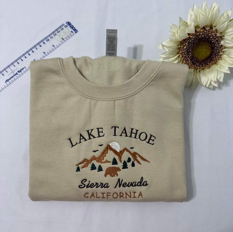 May include: A beige sweatshirt with an embroidered design of mountains, a bear, and trees. The text "LAKE TAHOE" is above the design, and the text "Sierra Nevada CALIFORNIA" is below the design.
