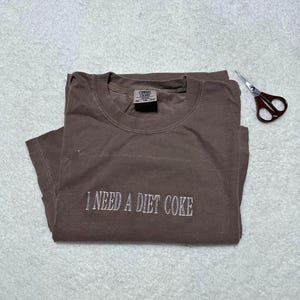 May include: A brown t-shirt with the text "I NEED A DIET COKE" embroidered in white on the front. The shirt is folded and laid flat on a white surface.