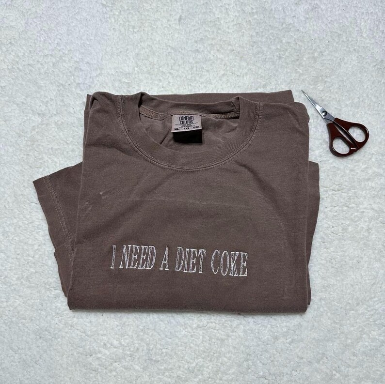 May include: A brown t-shirt with the text "I NEED A DIET COKE" embroidered in white on the front. The shirt is folded and laid flat on a white surface.