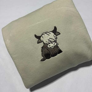 May include: A folded, light green sweatshirt featuring an embroidered design of a Highland cow covering its eyes. The cow is depicted in shades of brown and black, with the eyes and face in white. The sweatshirt is made of a soft material.