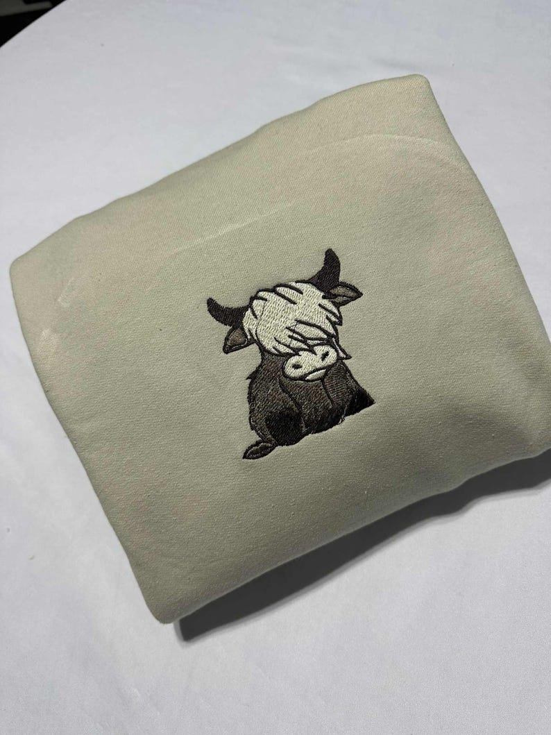 May include: A folded, light green sweatshirt featuring an embroidered design of a Highland cow covering its eyes. The cow is depicted in shades of brown and black, with the eyes and face in white. The sweatshirt is made of a soft material.