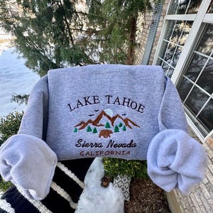 May include: Gray sweatshirt with embroidered text "LAKE TAHOE" and a mountain scene with a bear. The text "Sierra Nevada CALIFORNIA" is also embroidered below the mountain scene.