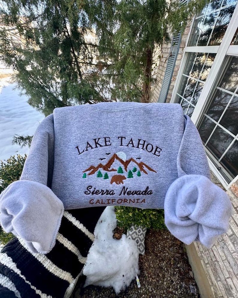 May include: Gray sweatshirt with embroidered text "LAKE TAHOE" and a mountain scene with a bear. The text "Sierra Nevada CALIFORNIA" is also embroidered below the mountain scene.