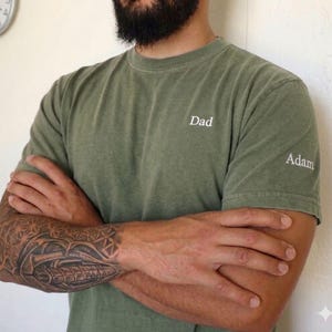 May include: Olive green t-shirt with the word "Dad" embroidered on the chest and "Adam" on the sleeve. The shirt is a solid color and has a classic crew neck design. The person's arms are crossed, revealing a detailed tattoo.