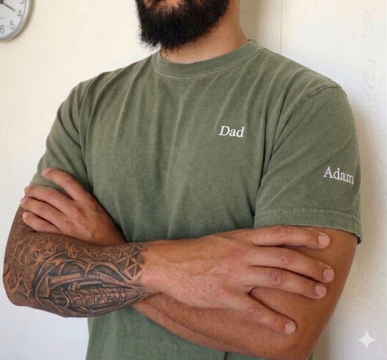 May include: Olive green t-shirt with the word "Dad" embroidered on the chest and "Adam" on the sleeve. The shirt is a solid color and has a classic crew neck design. The person's arms are crossed, revealing a detailed tattoo.