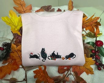 May include: A pink sweatshirt featuring an embroidered design of four black cats wearing Christmas hats and holding presents. The cats are arranged in a line across the front of the sweatshirt.