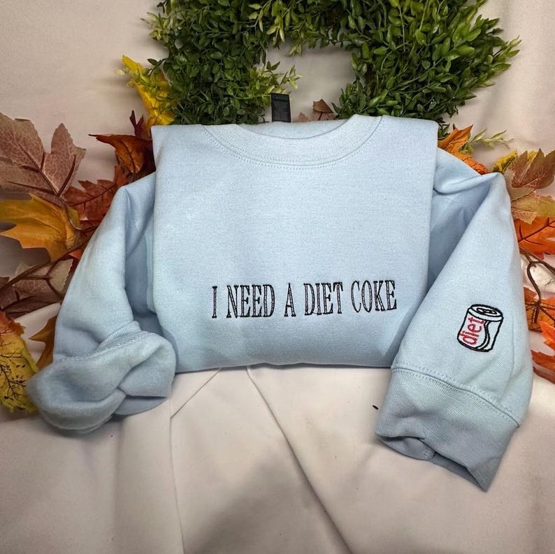May include: A light blue sweatshirt with the text "I need a Diet Coke" embroidered on the front and a small embroidered image of a Diet Coke can on the sleeve.