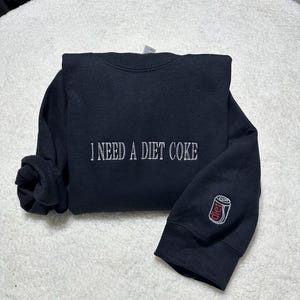 May include: Black sweatshirt with the text "I need a Diet Coke" embroidered on the front and a small embroidered image of a Diet Coke can on the sleeve.
