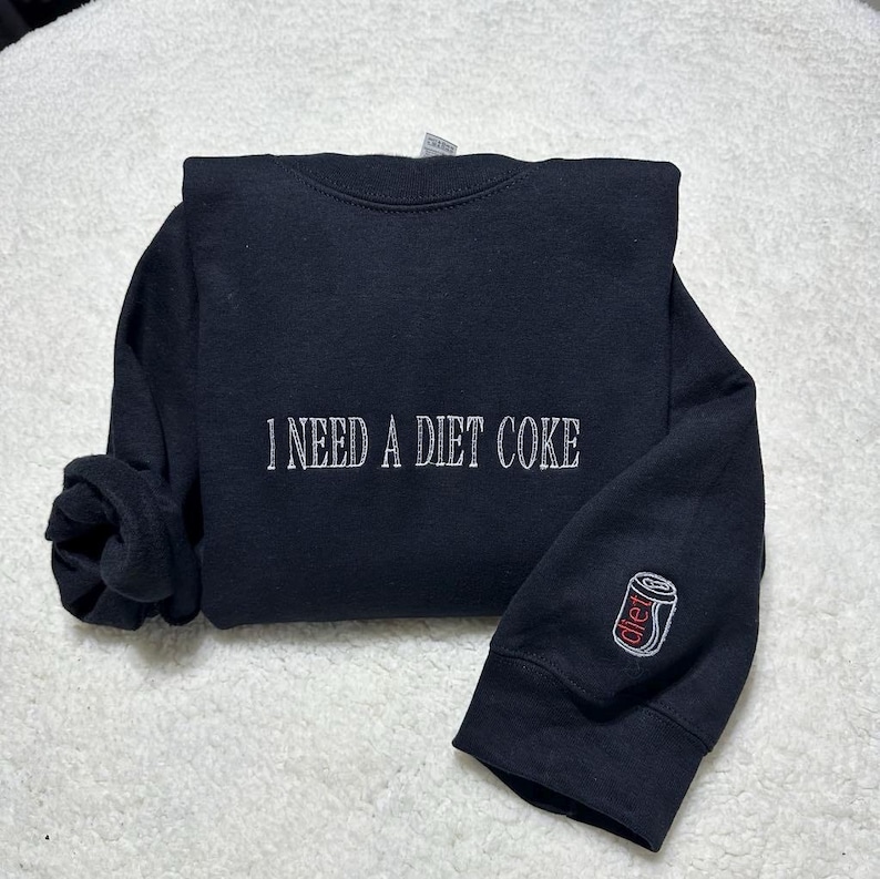 May include: Black sweatshirt with the text "I need a Diet Coke" embroidered on the front and a small embroidered image of a Diet Coke can on the sleeve.