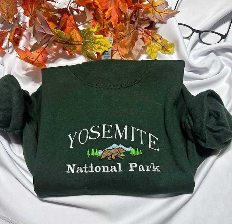 May include: A dark green sweatshirt with white embroidery that reads "Yosemite National Park". The embroidery also includes a brown bear and green trees.