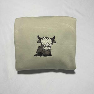 May include: A folded, light beige blanket featuring an embroidered Highland cow. The cow is depicted in black and brown with a white shaggy fringe. The blanket is folded neatly, showcasing the embroidered design.