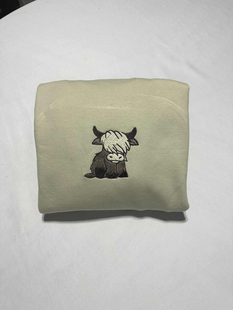 May include: A folded, light beige blanket featuring an embroidered Highland cow. The cow is depicted in black and brown with a white shaggy fringe. The blanket is folded neatly, showcasing the embroidered design.