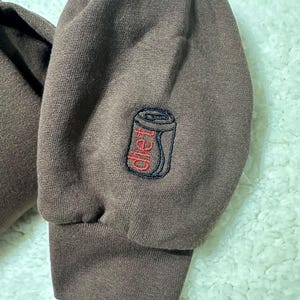 May include: Brown sweatshirt with a black and red embroidered image of a can of soda with the word "diet" on it.