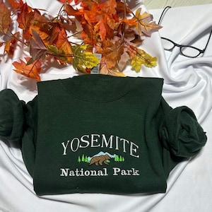 May include: A dark green sweatshirt with the text "Yosemite National Park" embroidered in white. The design includes a small embroidered image of a bear and a mountain range.