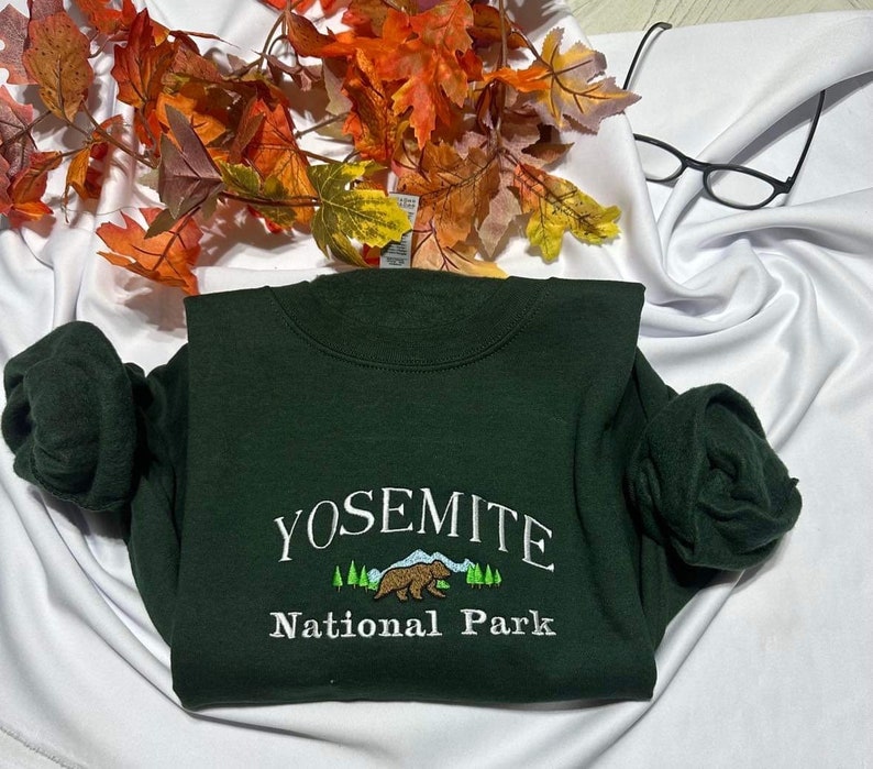 May include: A dark green sweatshirt with the text "Yosemite National Park" embroidered in white. The design includes a small embroidered image of a bear and a mountain range.