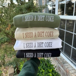 May include: Four stacked sweatshirts with the text "I NEED A DIET COKE" printed on each in white. The sweatshirts are in different colours: green, beige, white, and black.