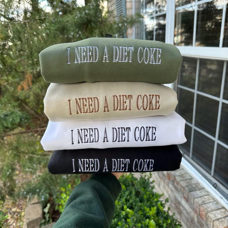May include: Four stacked sweatshirts with the text "I NEED A DIET COKE" printed on each in white. The sweatshirts are in different colours: green, beige, white, and black.
