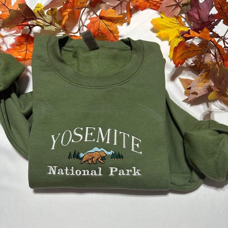 May include: A green sweatshirt with white embroidered text that reads "Yosemite National Park". The text is arched over a small embroidered image of a brown bear in a forest setting.