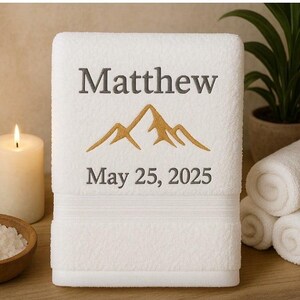 May include: White embroidered bath towel featuring the name "Matthew", a gold mountain design, and the date "May 25, 2025". The towel is displayed with rolled towels, a lit candle, and a bowl of salt.