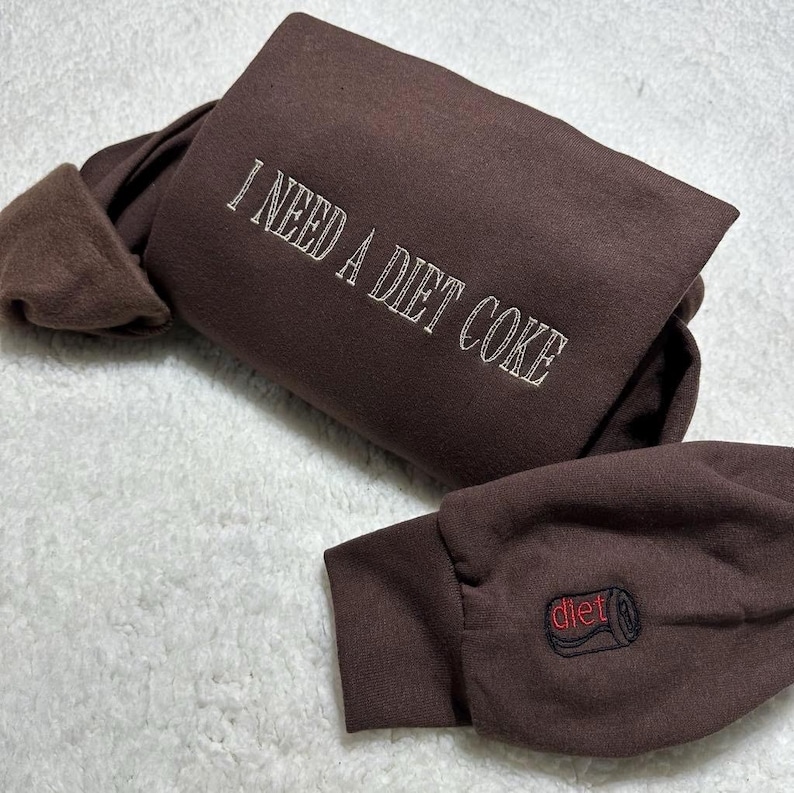 May include: Brown sweatshirt with the text "I need a diet coke" embroidered on the front and a red "diet" can logo on the sleeve.